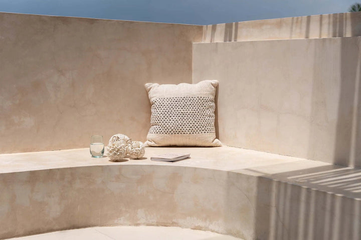 Beige stone bench with a textured pillow and decorative items on a sunny day.