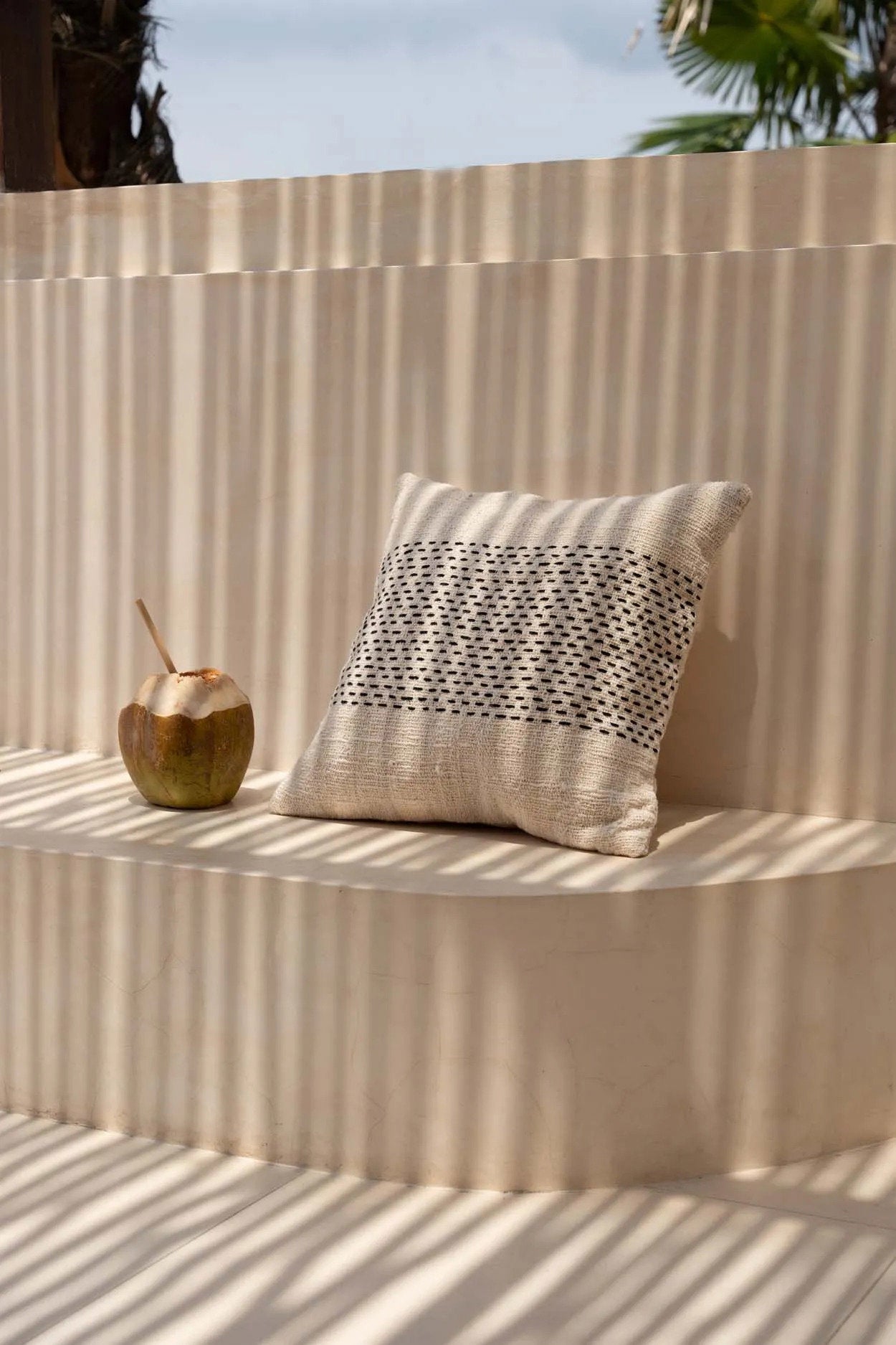Decorative pillow with a geometric pattern on a textured wall with a coconut and palm leaves in the background.