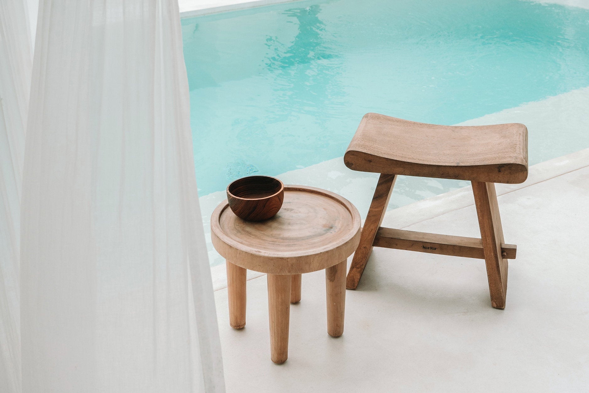 Wooden stool and small round table by a poolside with a white curtain.