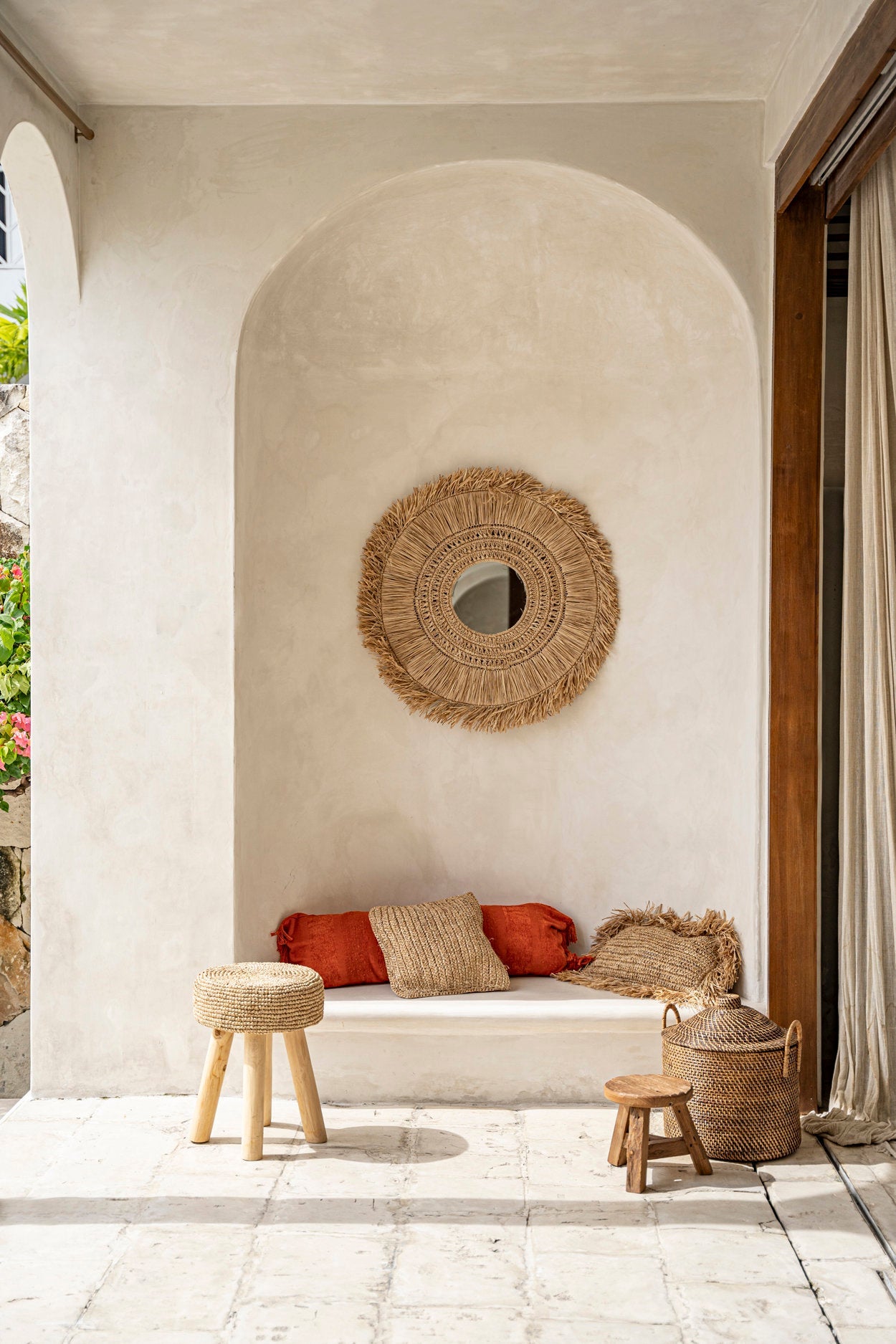 Outdoor seating area with a woven mirror, bench, and stools against a light-colored wall.