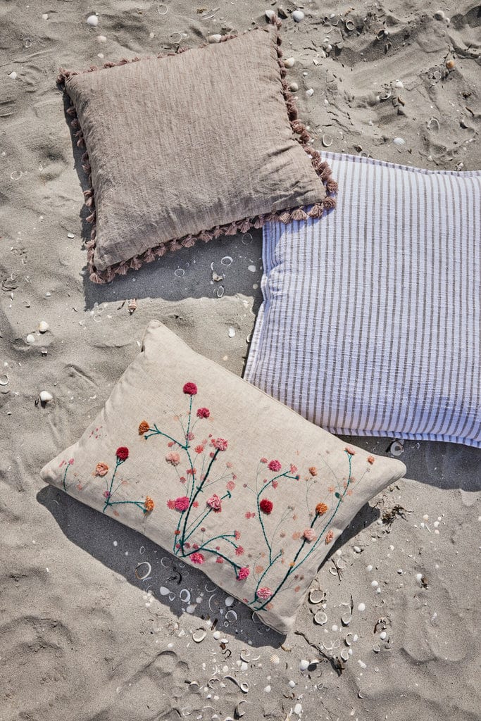 Oville Cushion 50x50 cm Dark Linen with tassels on sandy beach background.