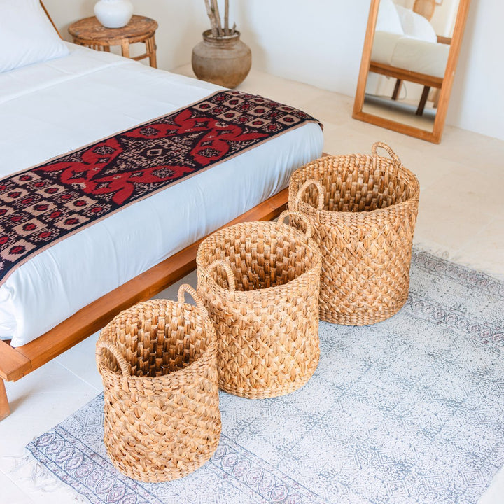 Three woven baskets on a rug in a bedroom setting with a bed and mirror in the background.