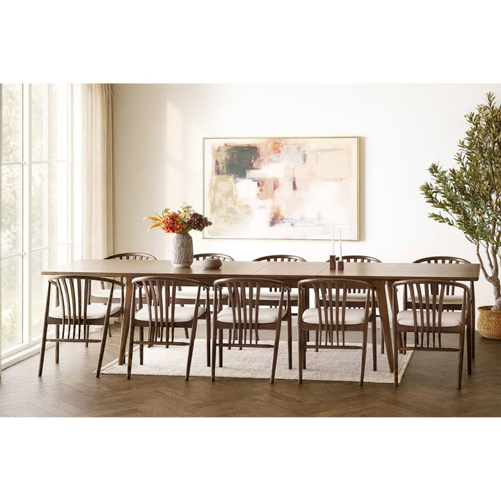 A dining room scene showing a rectangular wooden dining table with ten matching slatted-back chairs, set on a light rug in a bright interior with a large abstract painting on the wall and a potted tree.