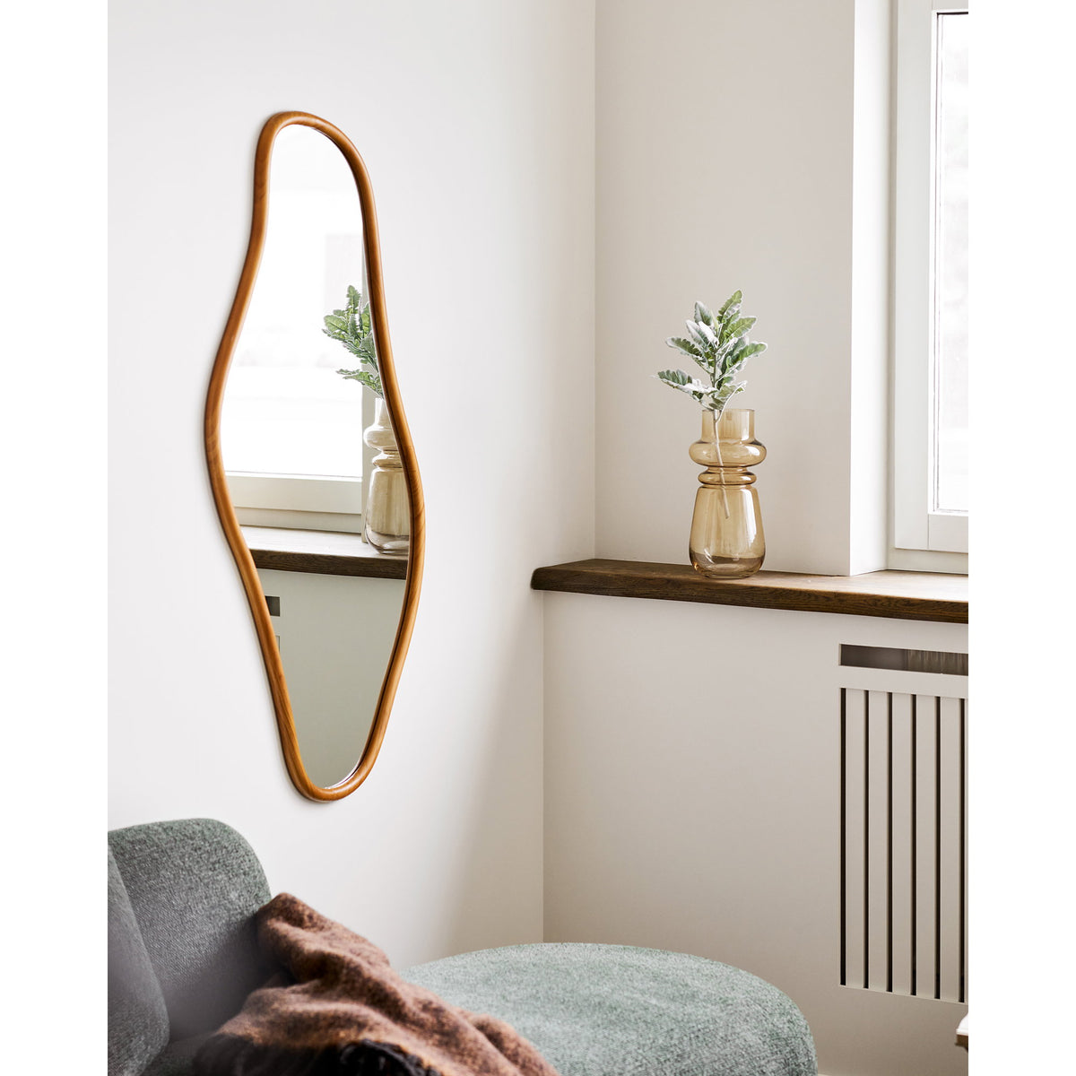 Oval mirror with wooden frame on a wall above a sofa with a plant on a windowsill.