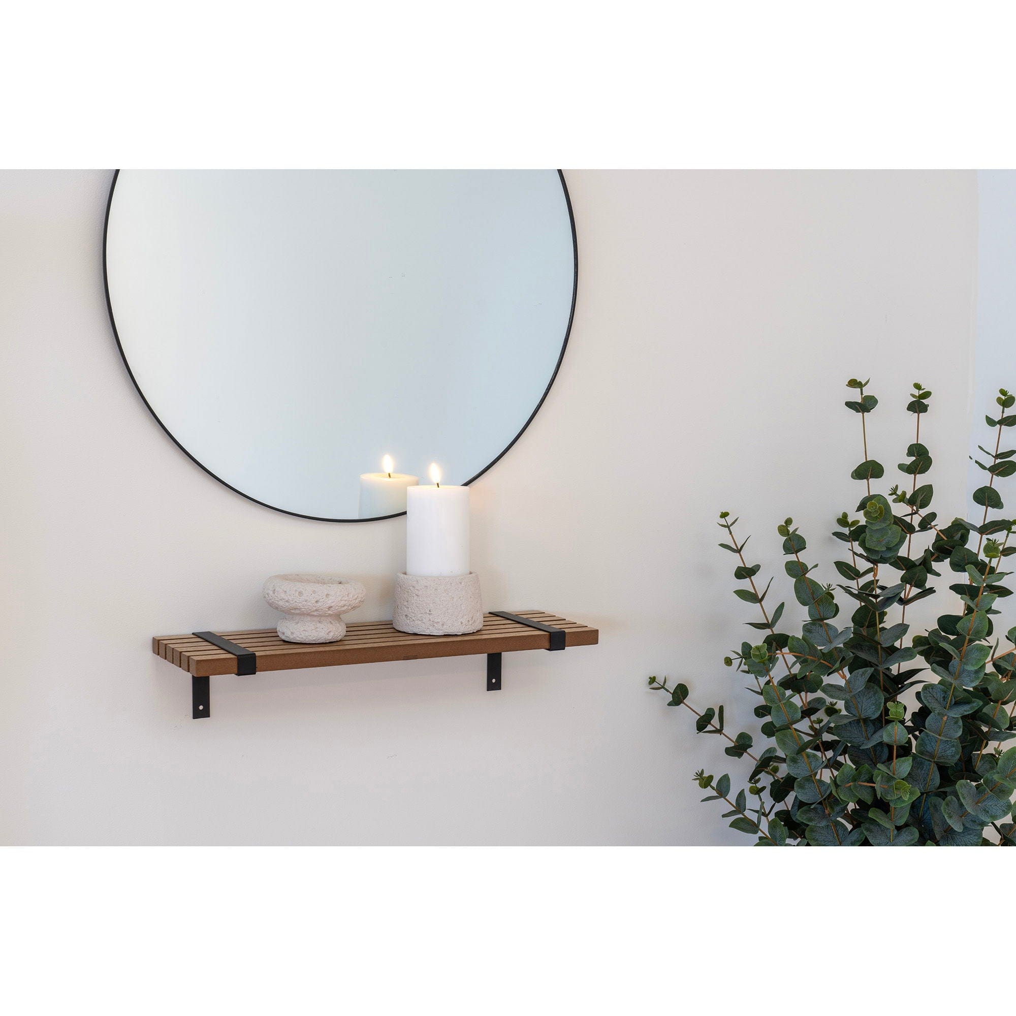 Round mirror above a wooden shelf with a candle and decorative objects, next to a potted plant.