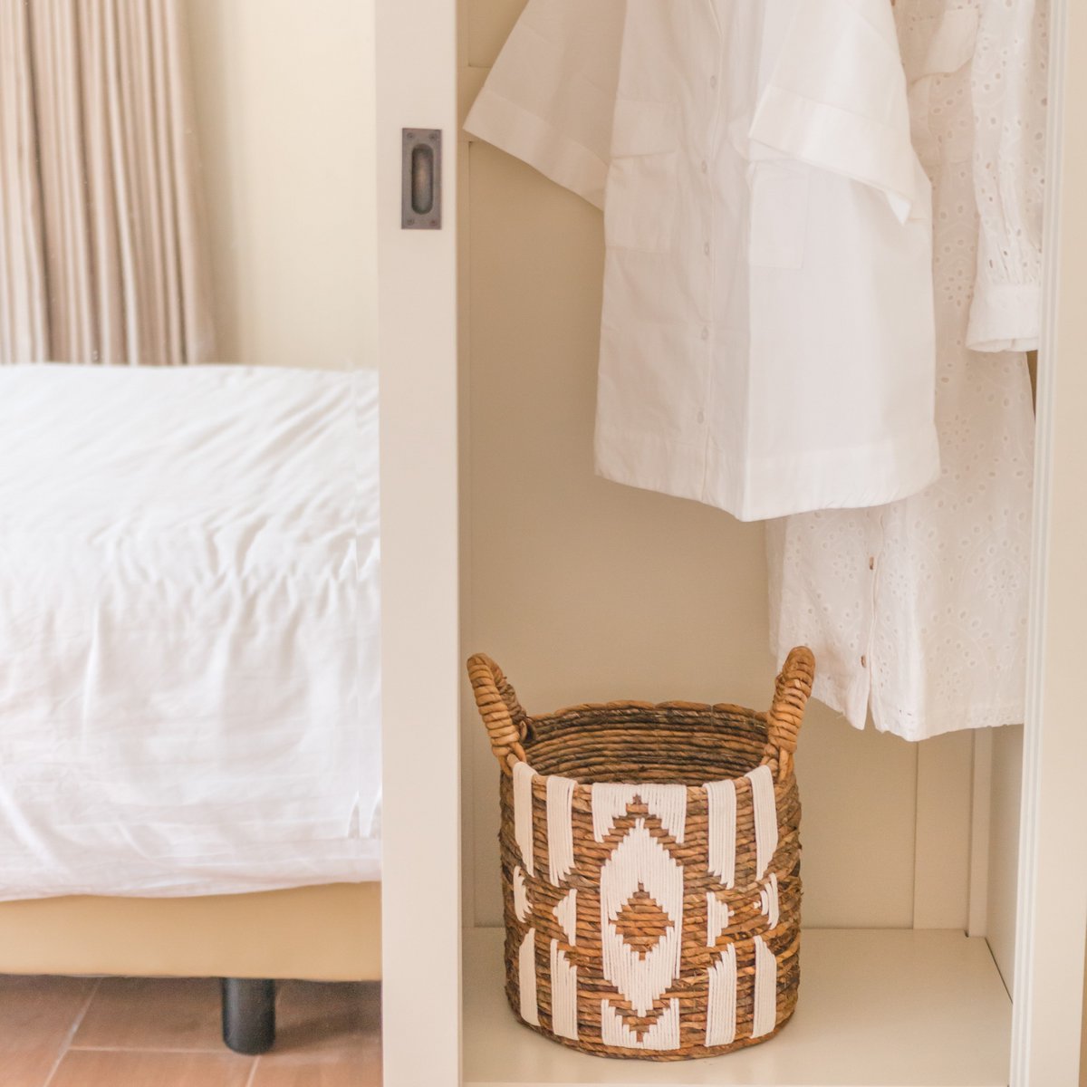 Woven basket with geometric pattern on a shelf next to a bed and hanging clothes.