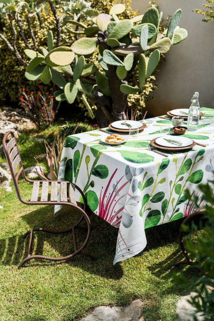 Outdoor setting with a table set for a meal on a green leaf-patterned tablecloth, surrounded by cacti and plants.