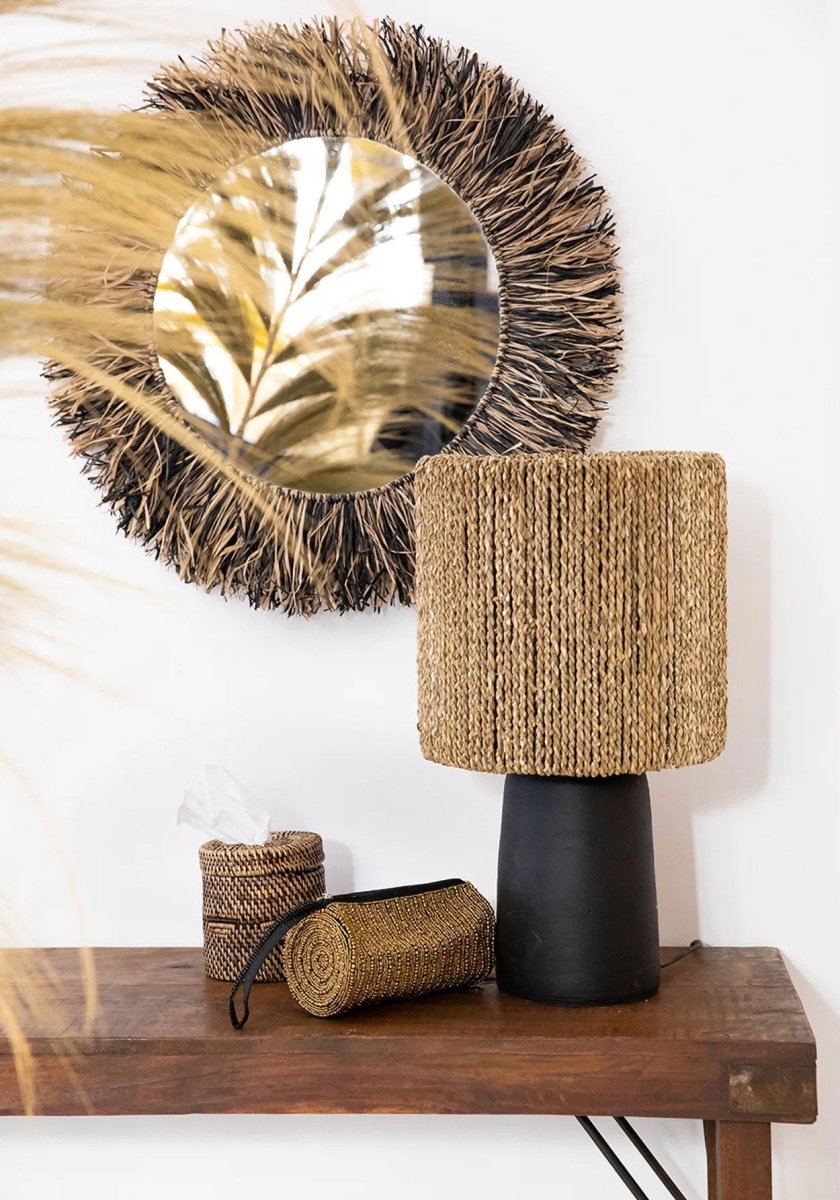 Decorative items including a woven mirror, lamp, and cylindrical containers on a wooden surface.