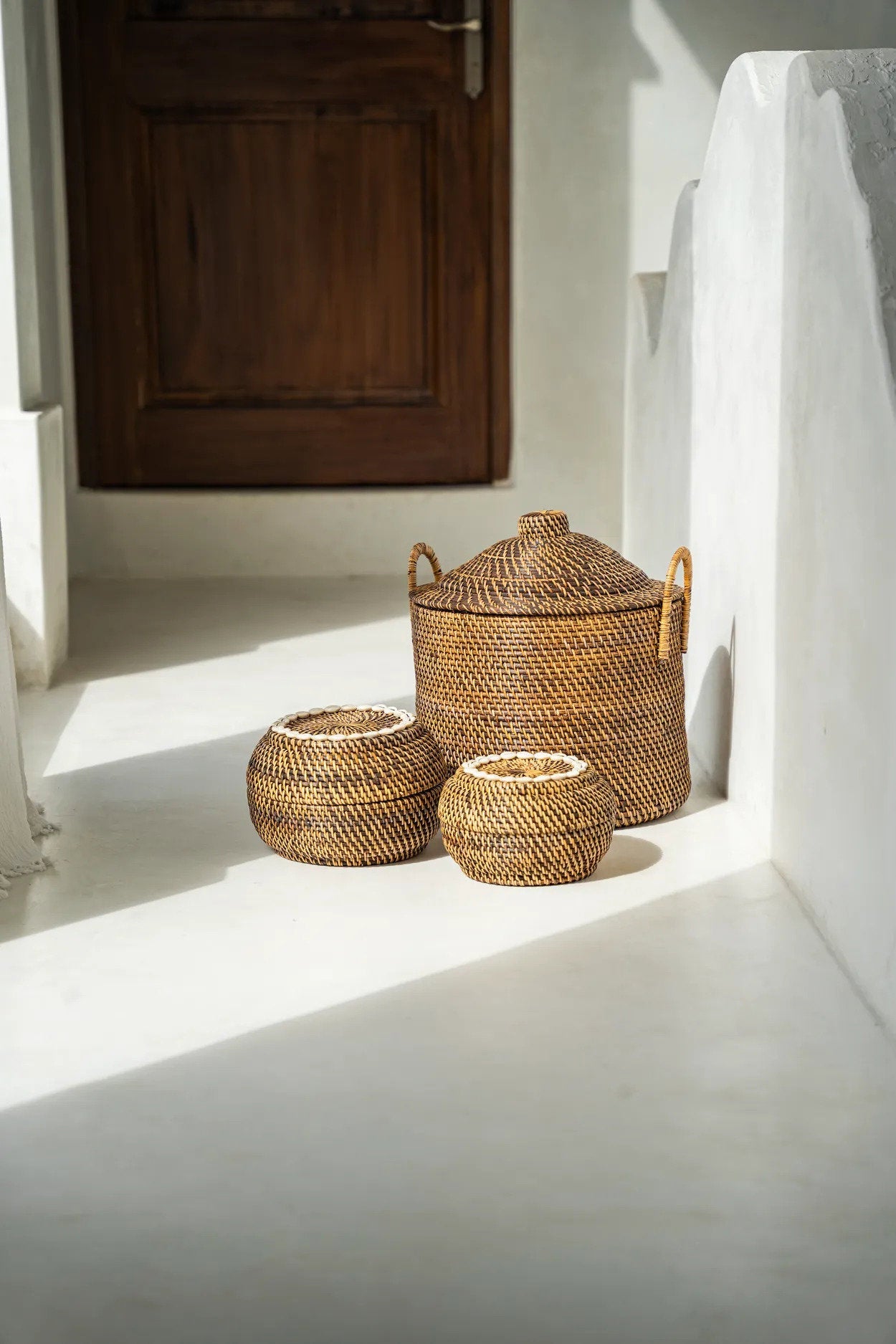 Three woven baskets with lids on a white floor with sunlight casting shadows.