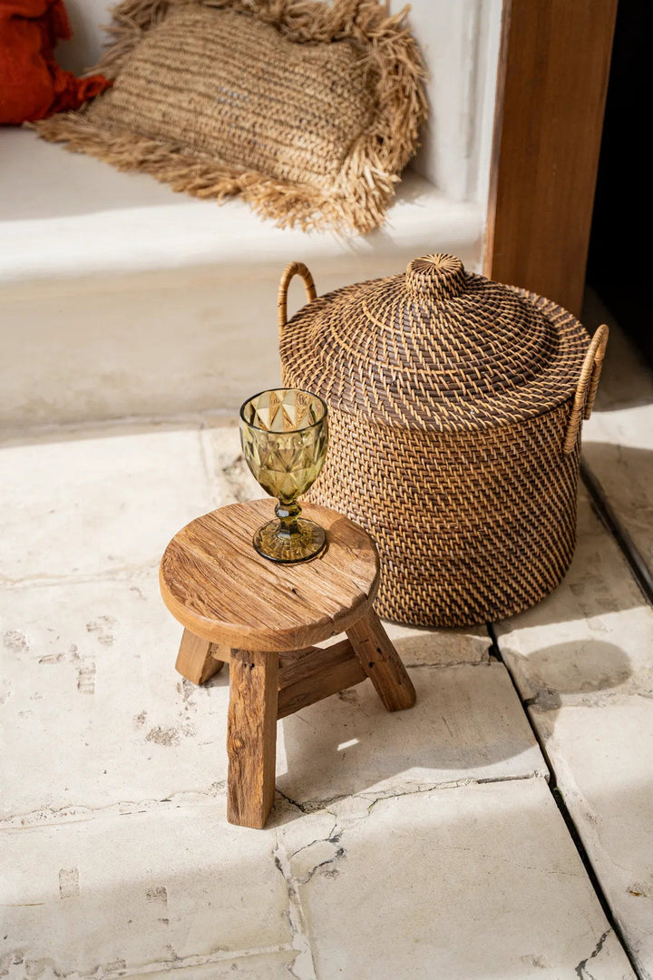 Woven basket, small wooden stool with a glass, and textured pillows on a stone floor.