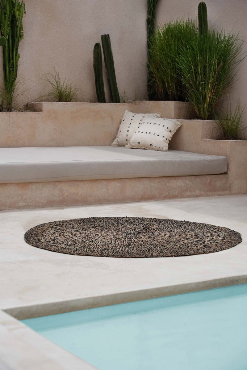Outdoor setting with a round woven mat, cushioned bench, and cacti near a pool.