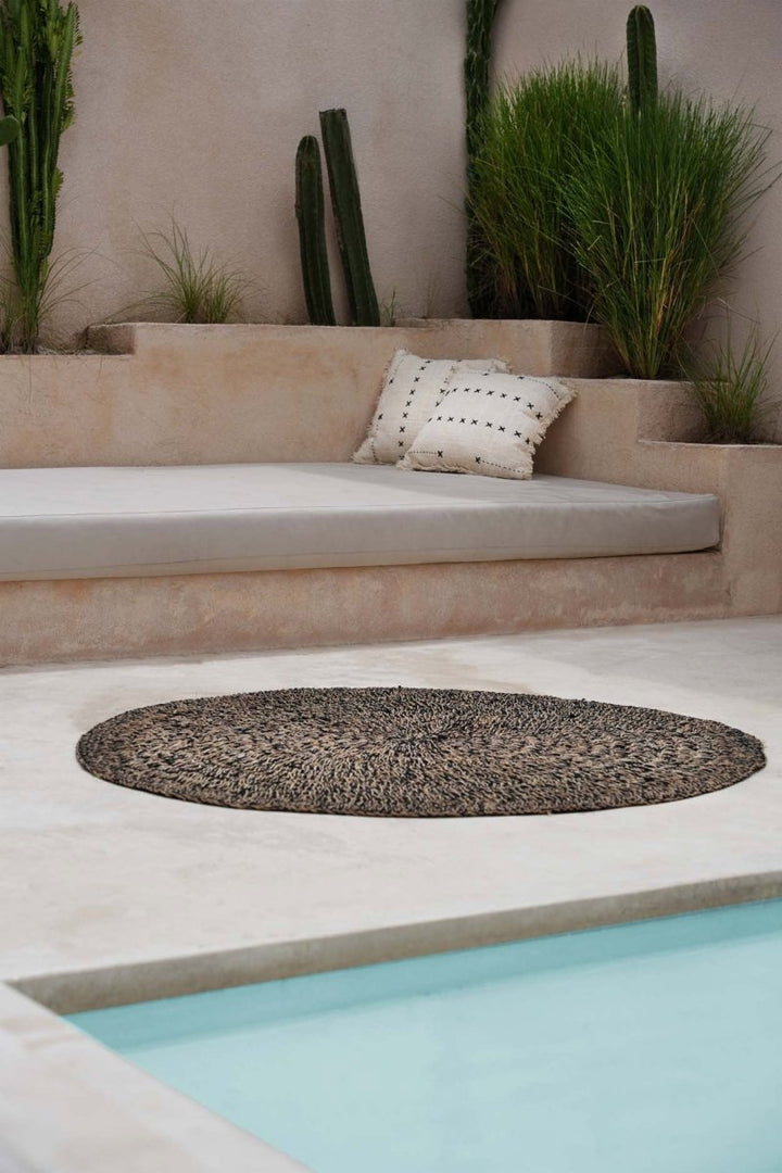 Outdoor setting with a round woven mat, cushioned bench, and cacti near a pool.