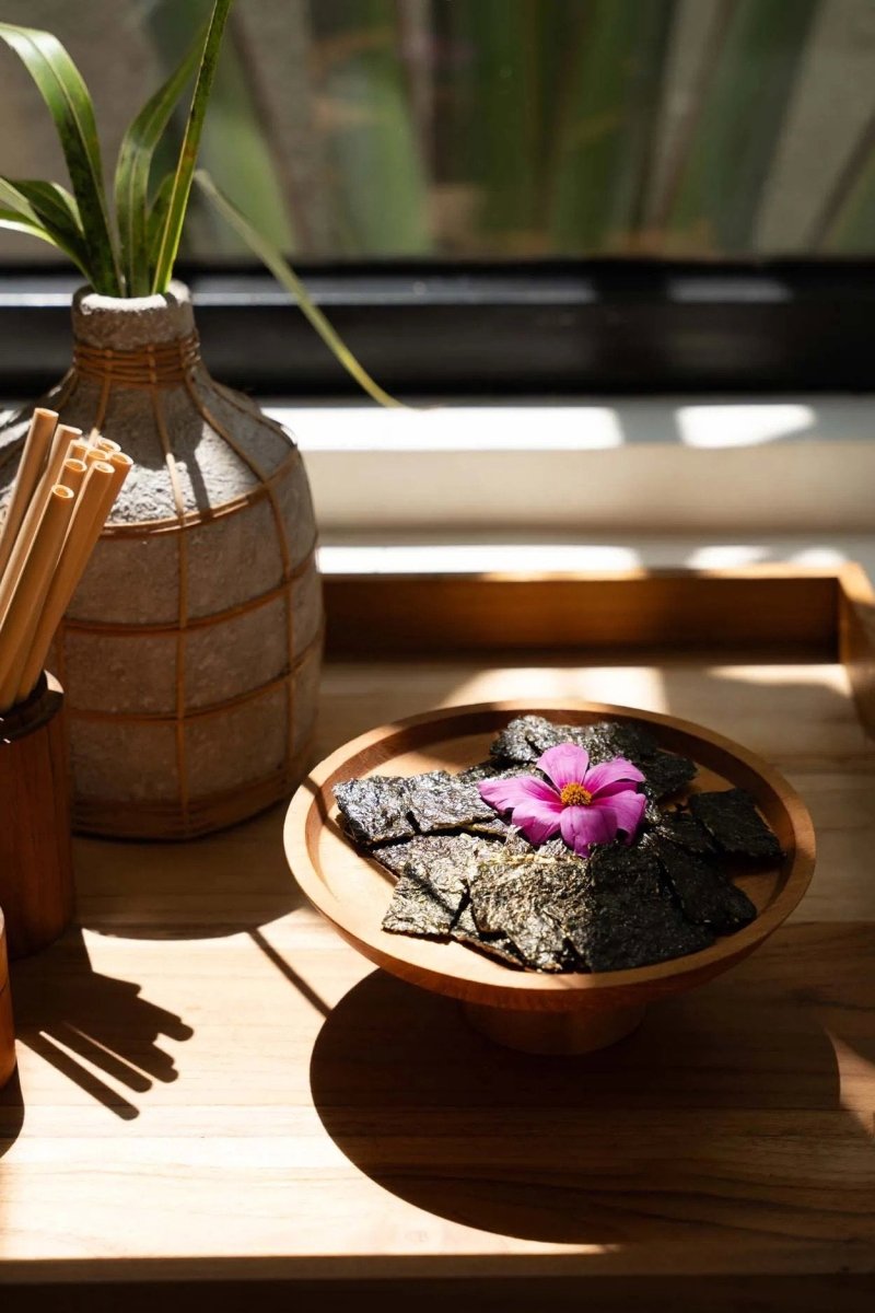 Bowl of seaweed with a pink flower on a wooden surface, next to a vase with a plant.