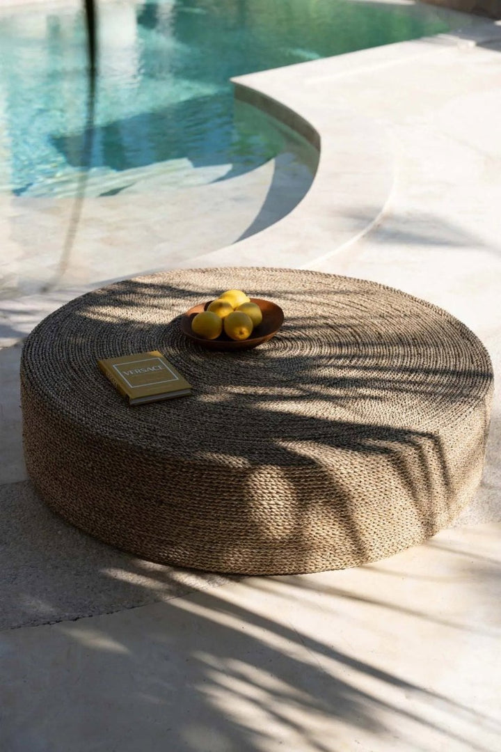 Round woven ottoman with a small tray of lemons by a poolside.