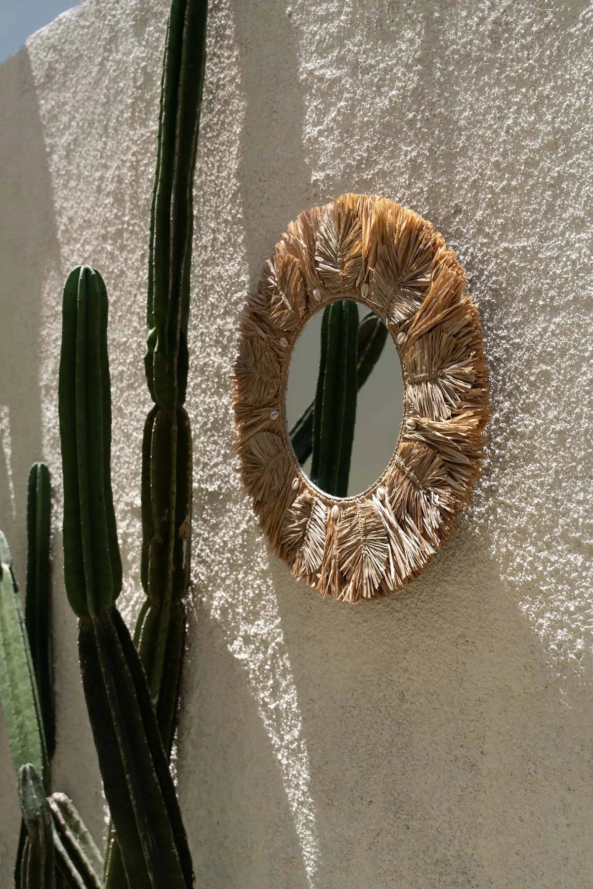 Round woven mirror on a textured wall with cacti in the foreground
