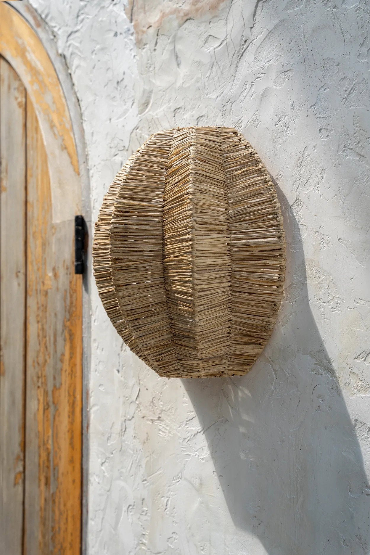 Round woven wall lamp hanging on a textured white wall next to a wooden door.