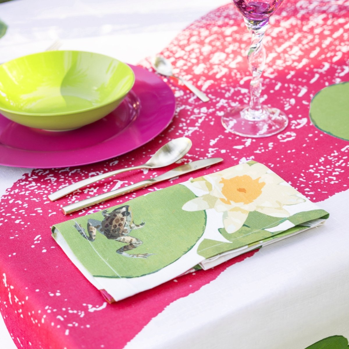Table setting with a pink tablecloth, green bowl, purple plate, floral napkin, and glass.