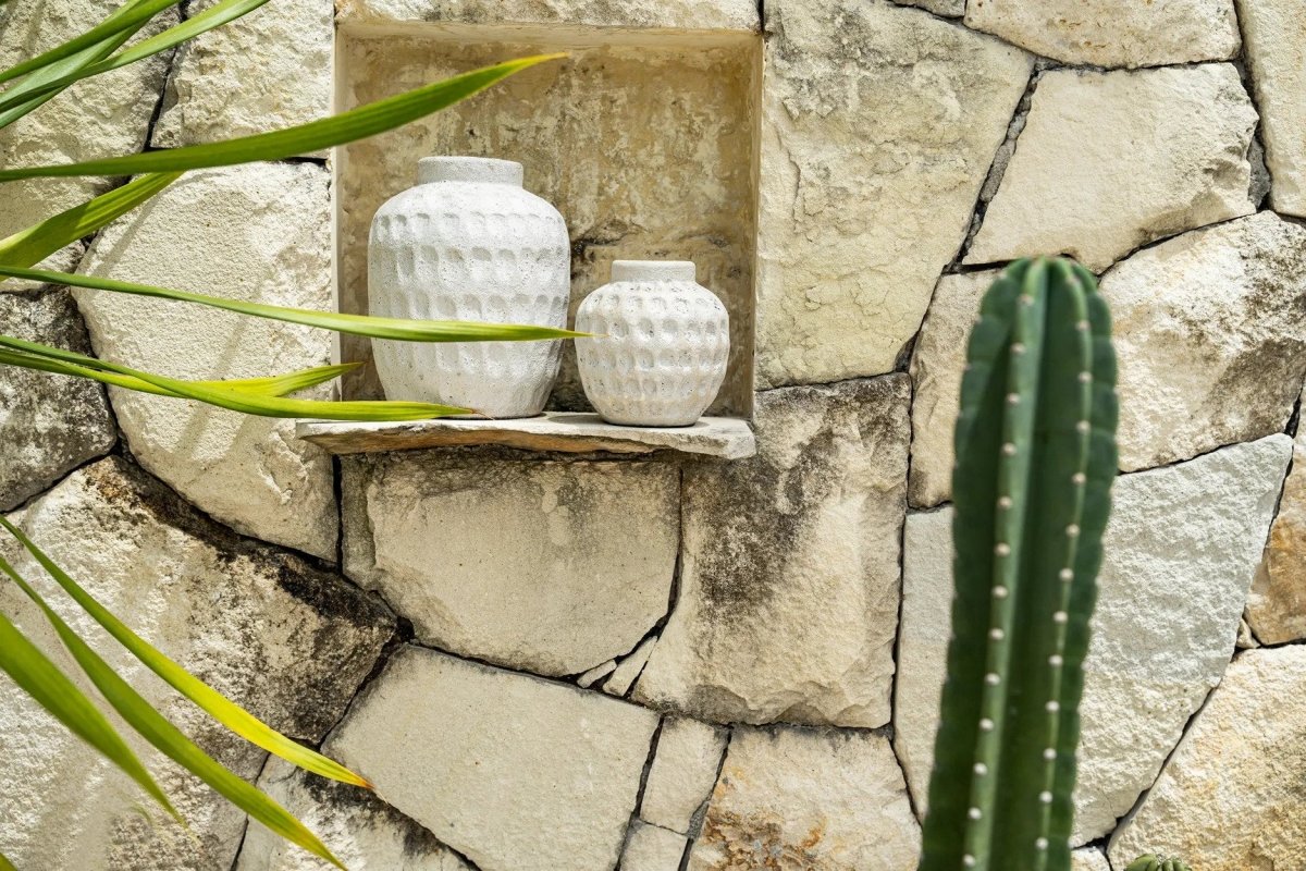 Two white textured vases on a stone shelf with a stone wall and plant in the background