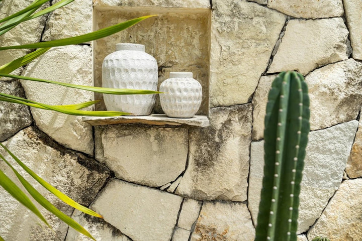 Two white textured vases on a stone shelf with a stone wall and plant in the background