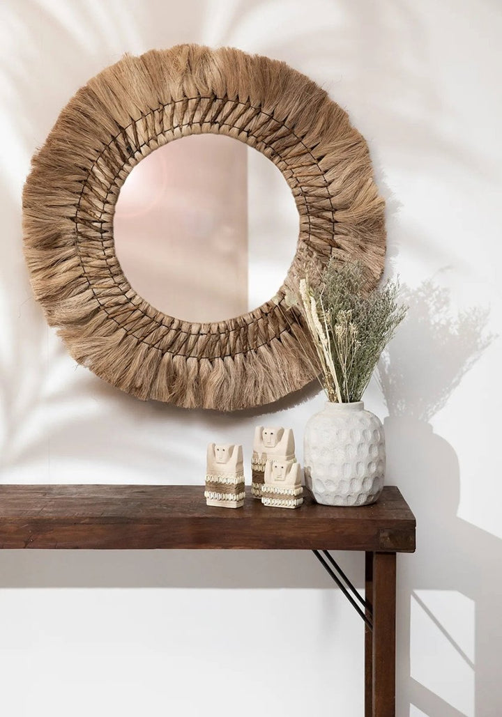 Round woven mirror on a wall above a wooden console table with decorative items.