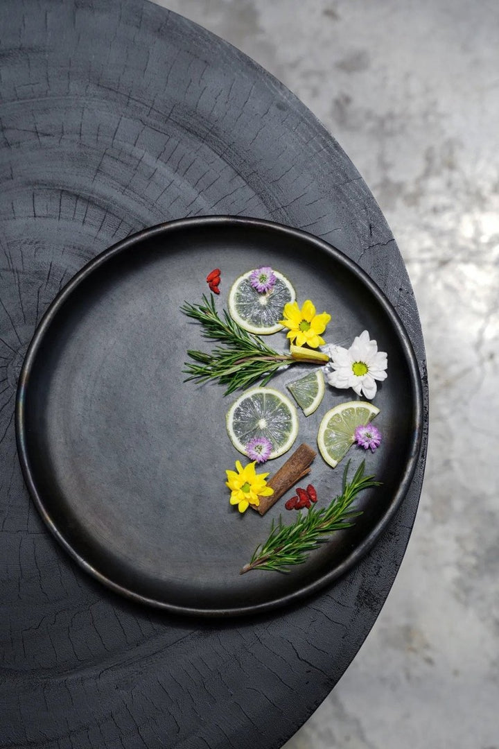Decorative arrangement of herbs, flowers, and citrus on a black plate with a textured surface.
