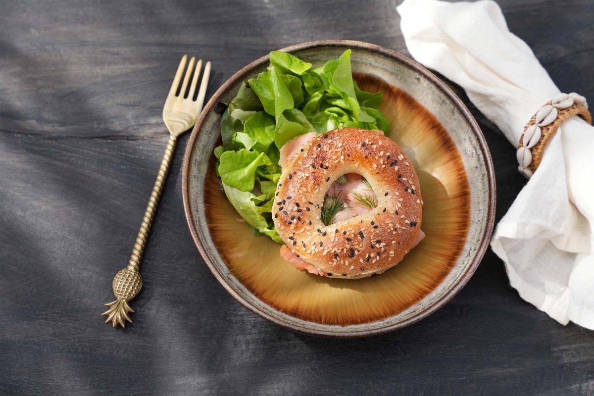 Bagel with lox and salad on a plate with a fork and napkin on a dark surface