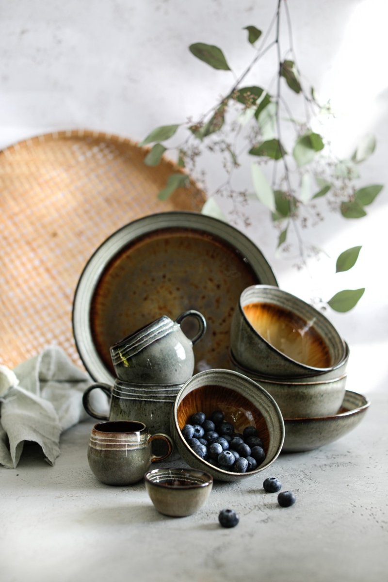 Set of ceramic bowls and mugs on a light surface with a woven basket and greenery in the background.