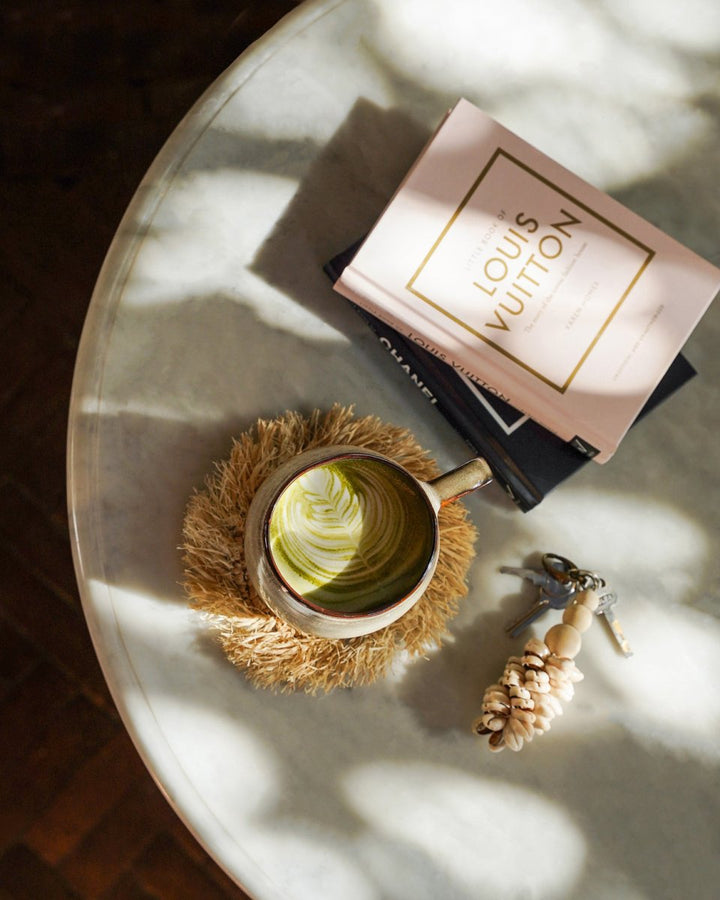 Cup of coffee on a textured coaster with a Louis Vuitton book and keys on a marble surface.
