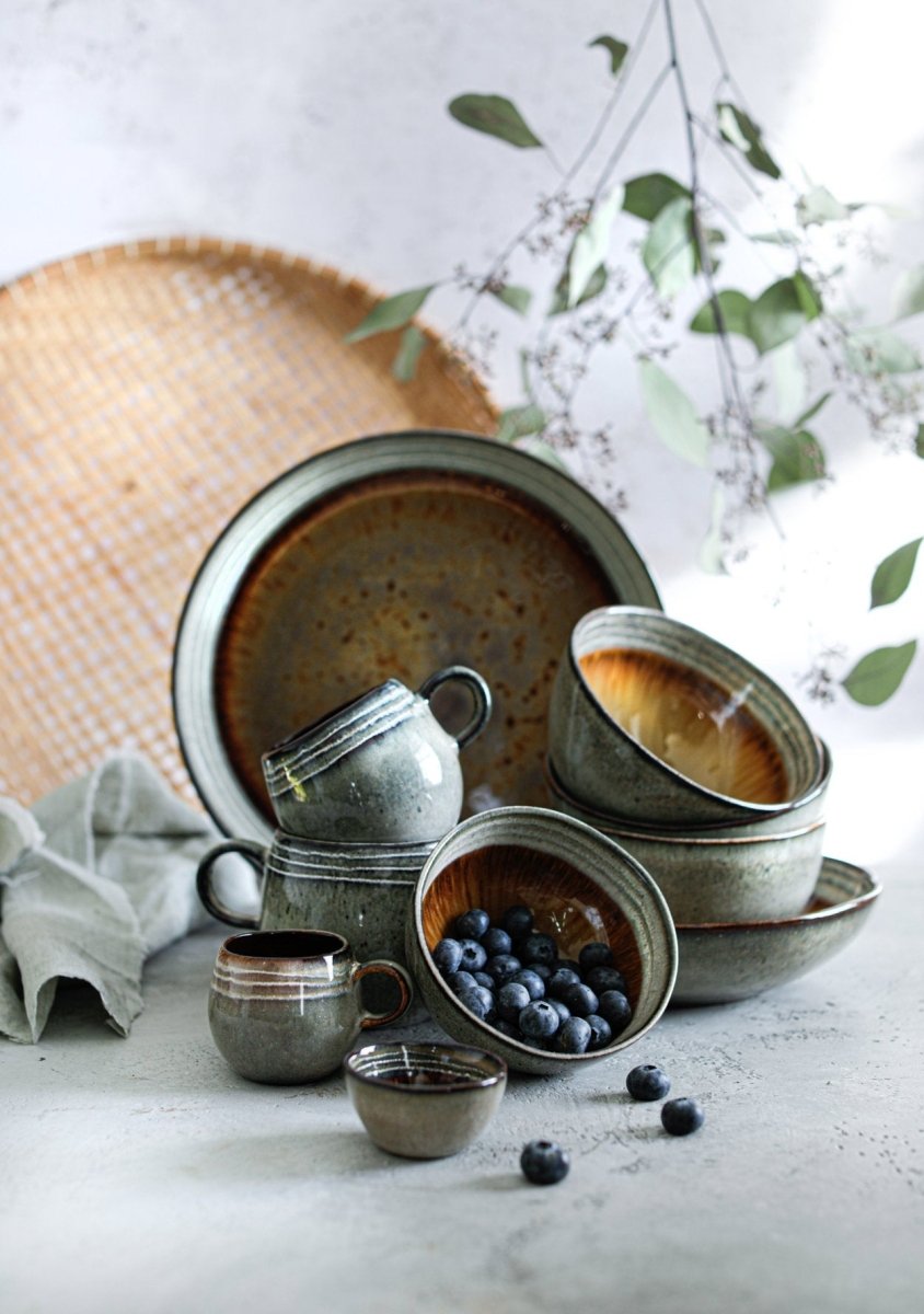 Set of ceramic bowls and cups on a light surface with a woven basket and greenery in the background.