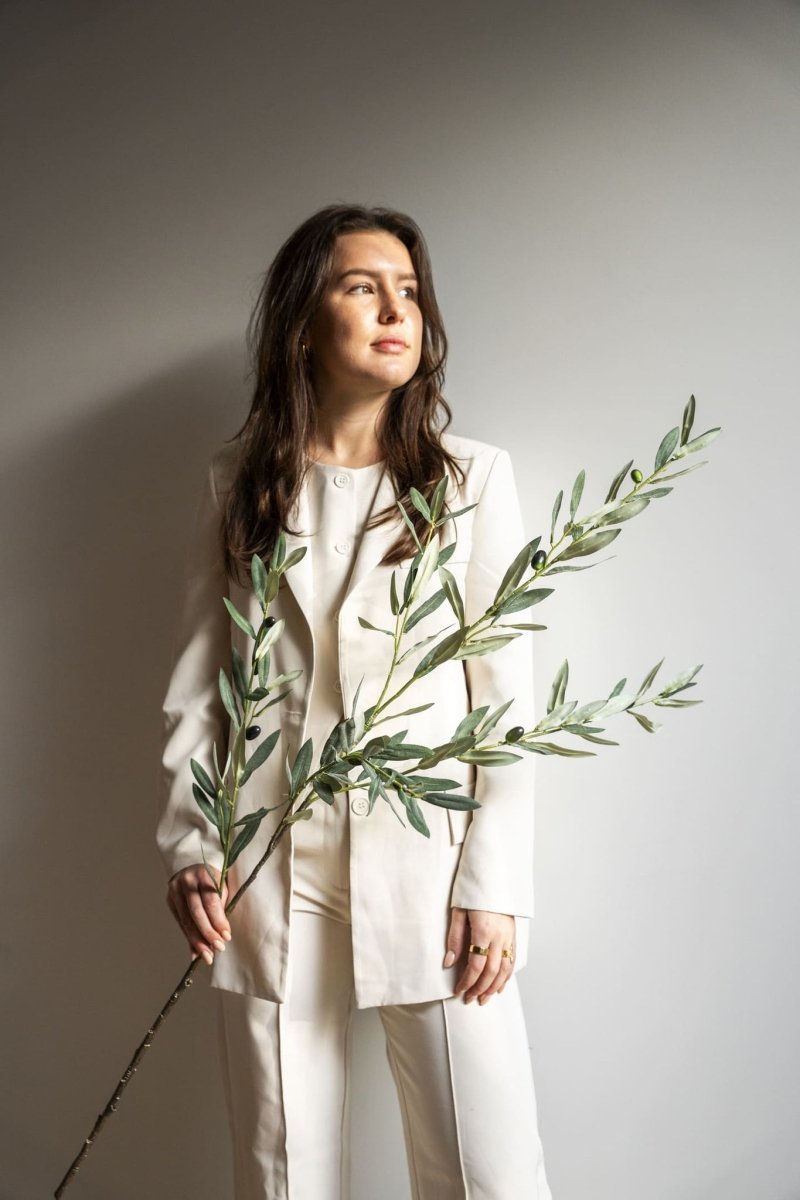 Woman in a white suit holding an olive branch against a plain background