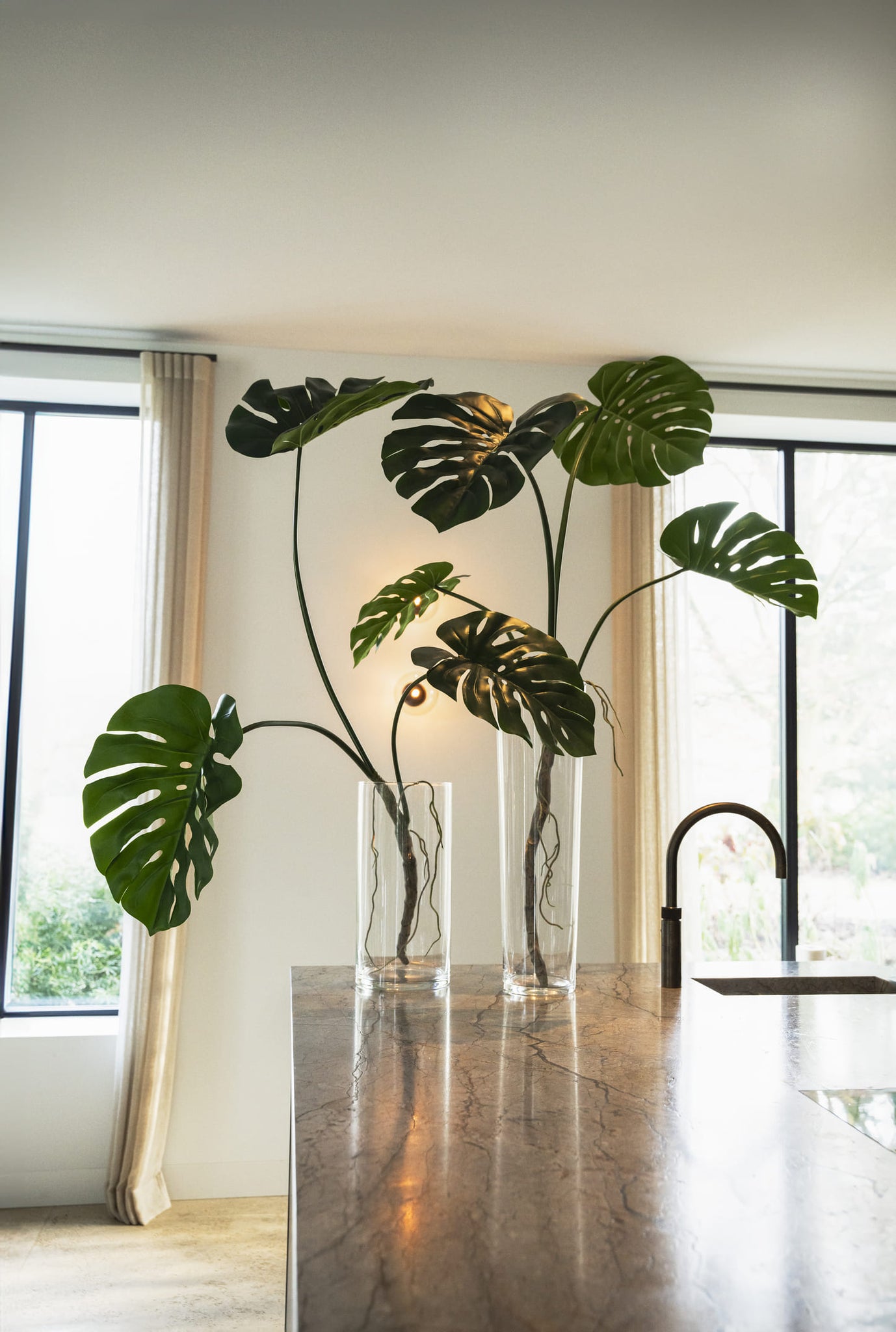 Monstera plants in clear vases on a marble surface with large windows in the background