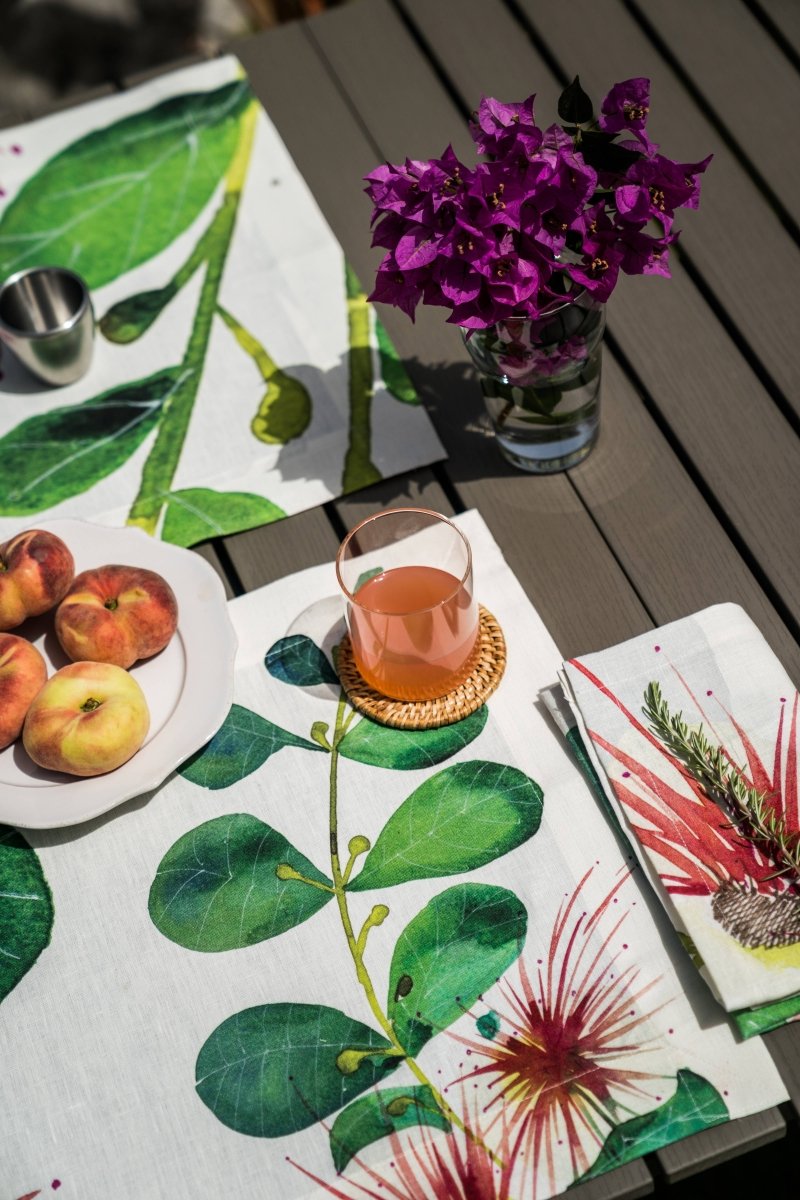 Table setting with floral tablecloth, peaches, glass of juice, and purple flowers.