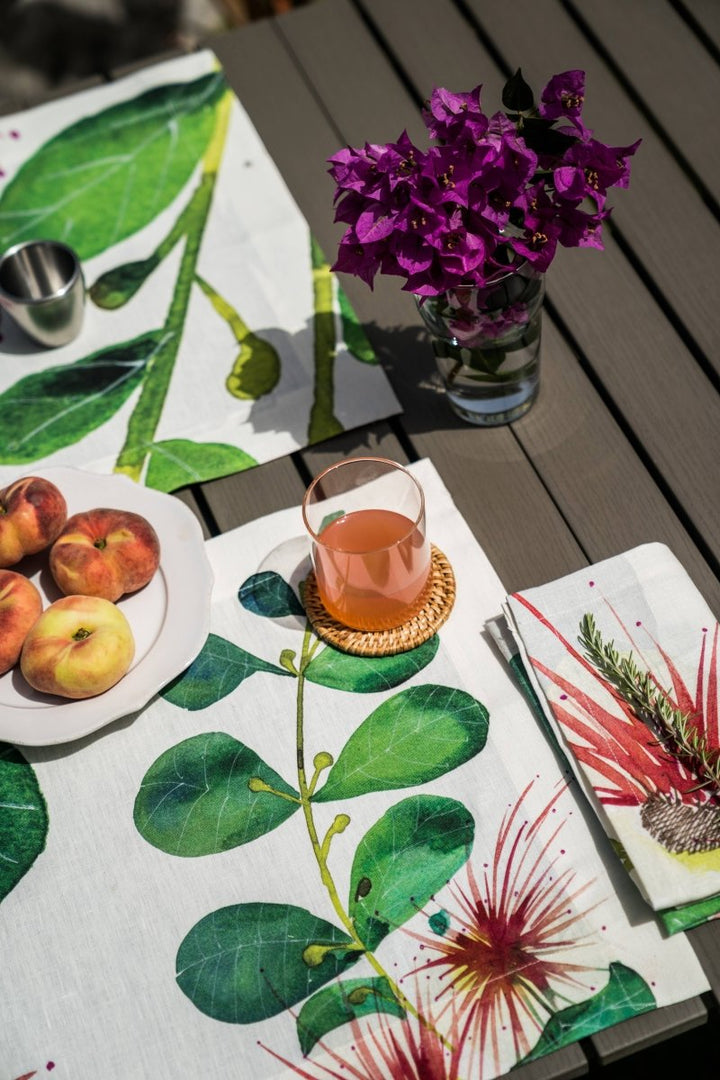 Table setting with floral tablecloth, peaches, glass of juice, and purple flowers.