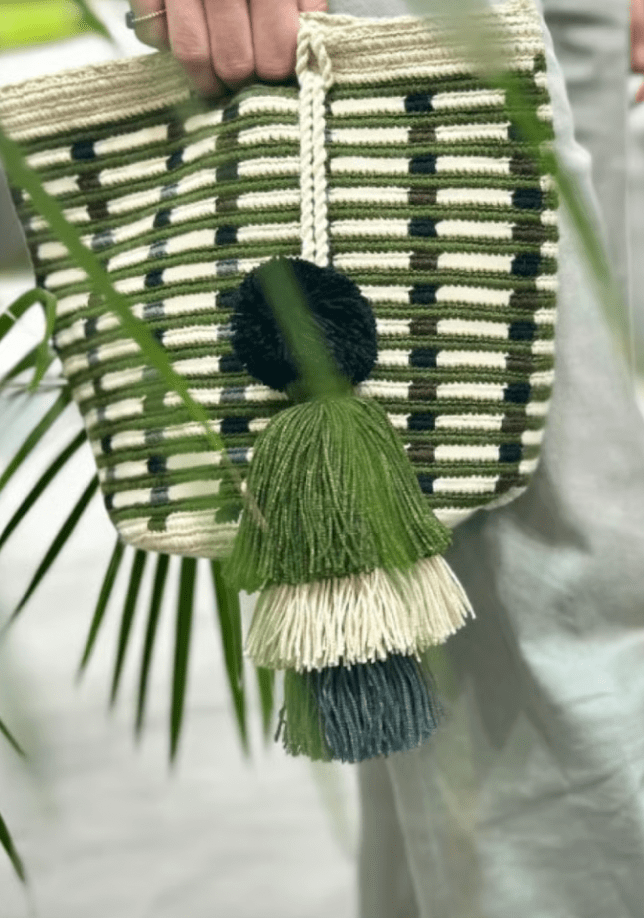 Handbag with green and white striped pattern and tassels held by a person against a blurred natural background.